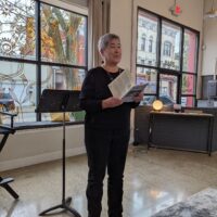 Gallery 2 - A woman with short grey hair reading a poem from a book.