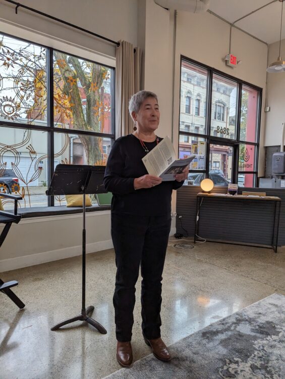 Gallery 2 - A woman with short grey hair reading a poem from a book.